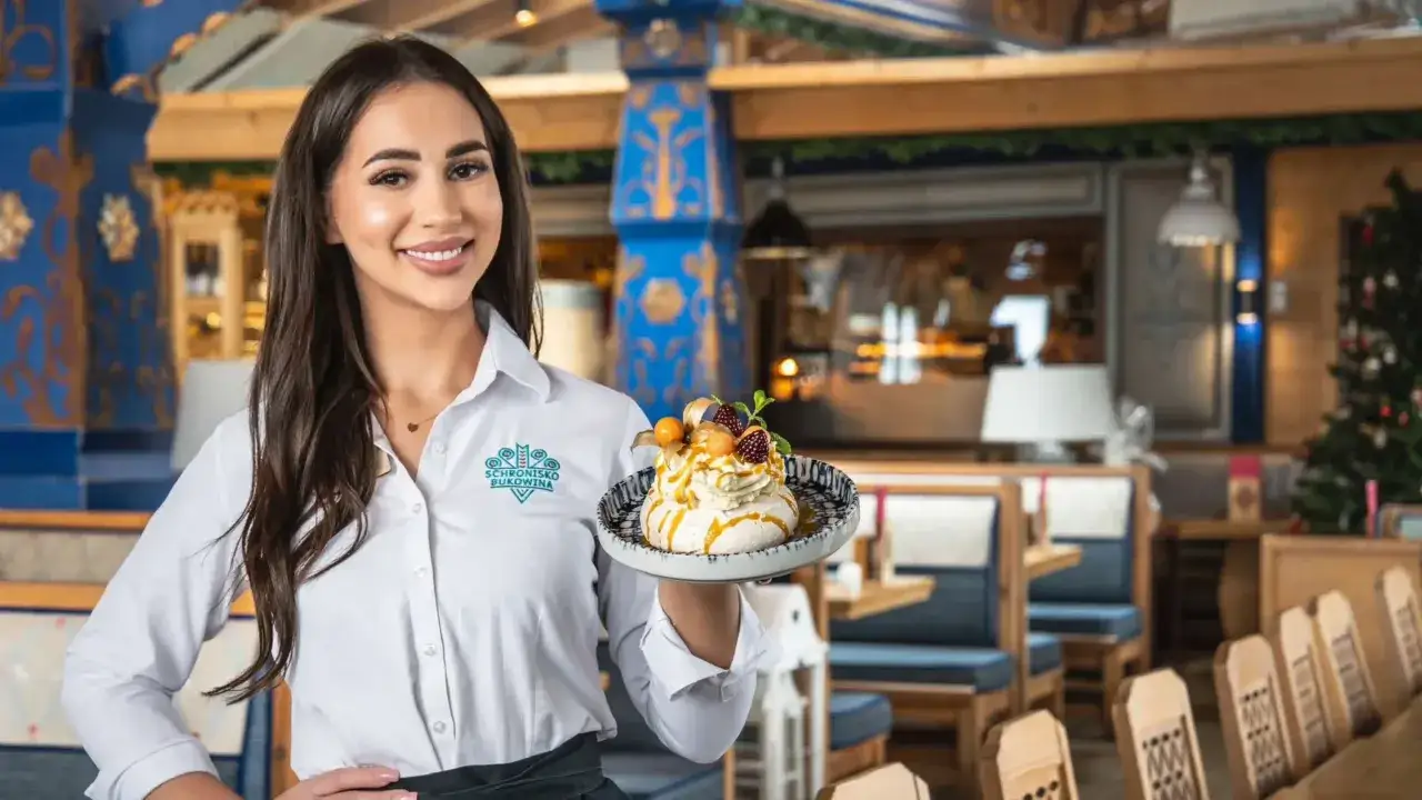Kelnerka prezentuje deser z menu Schroniska Smaków Bukowina. Wnętrze restauracji zdobią góralskie wzory.