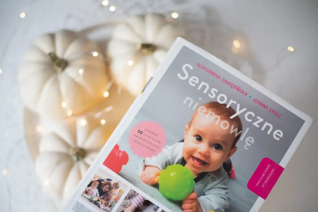 Okładka książki "Sensoryczne niemowlę" z uśmiechniętym dzieckiem, dyniami i lampkami.