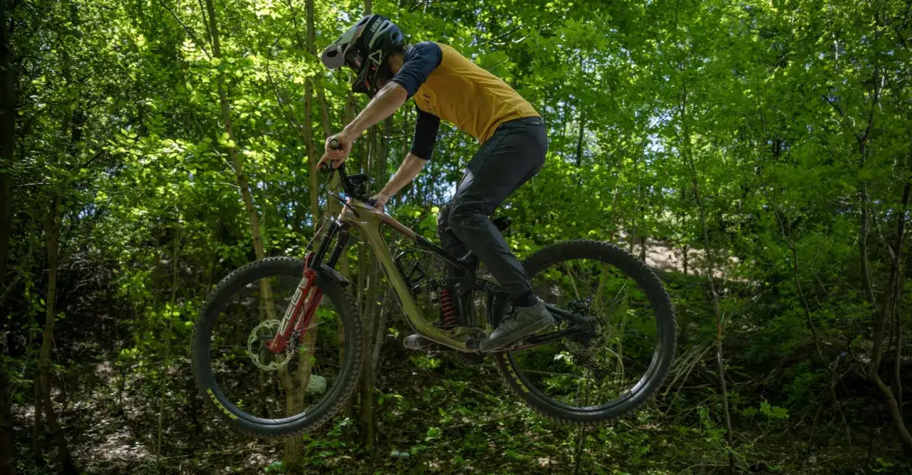 Kolarz w kasku wykonuje efektowne hopy rowerowe, unosząc się w powietrzu pośród zielonego lasu.
