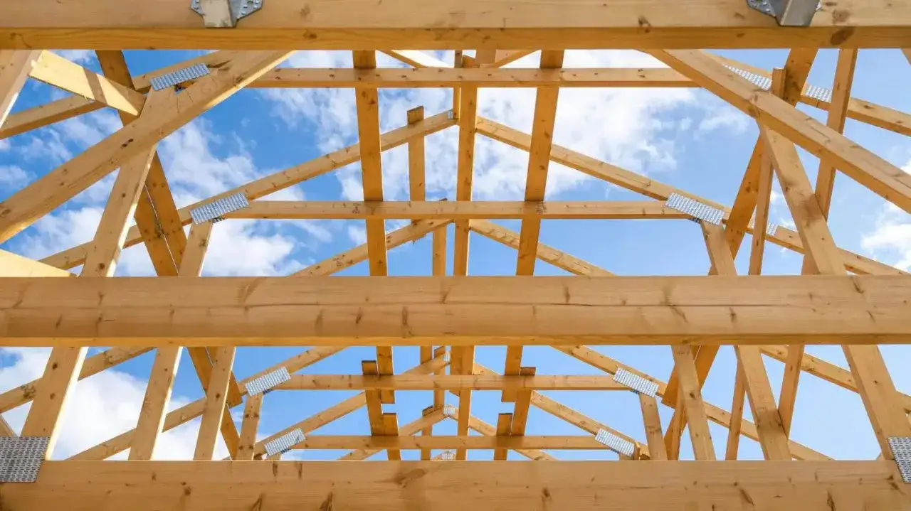 Drewniane wiązary tworzą solidną więźbę dachową, gotową na przyjęcie pokrycia. Widać błękitne niebo z chmurami.