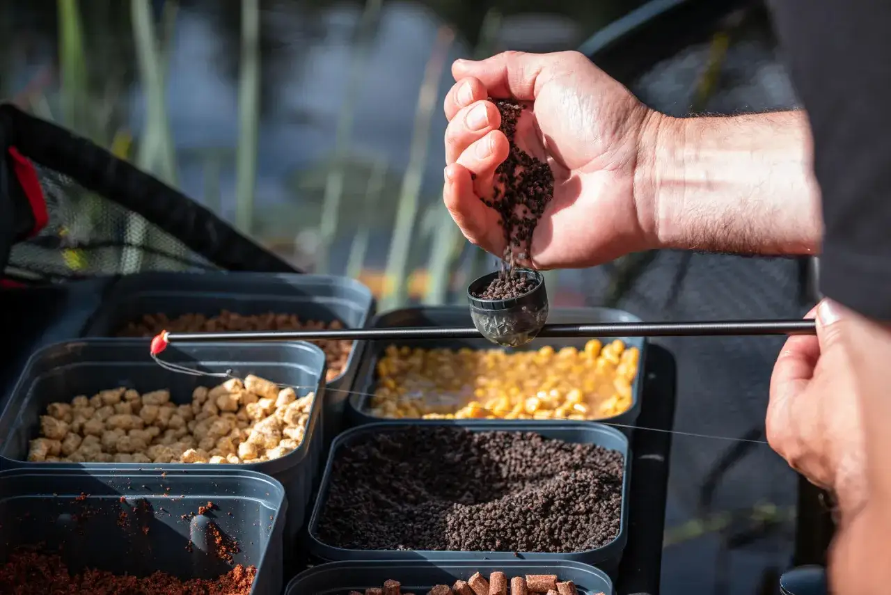 Dłoń wsypuje zanętę do małego pojemnika na tle pojemników z różnymi przynętami wędkarskimi.