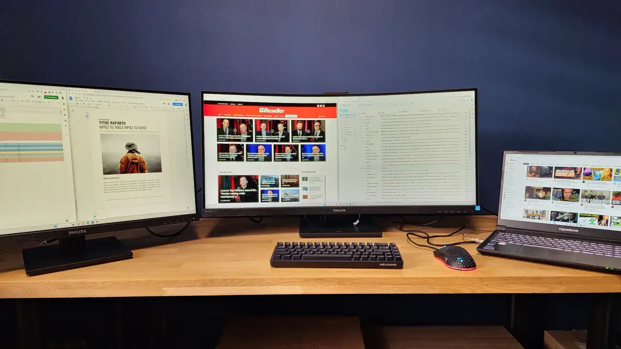 Dwa monitory i laptop z otwartymi oknami, klawiatura i myszka na drewnianym biurku.