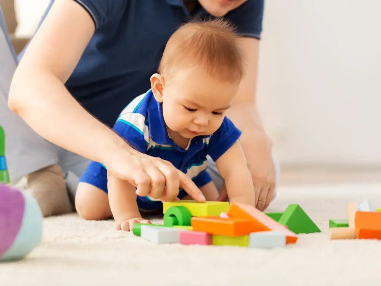 Dziecko w niebieskiej koszulce bawi się kolorowymi klockami na dywanie, a obok niego siedzi dorosły.