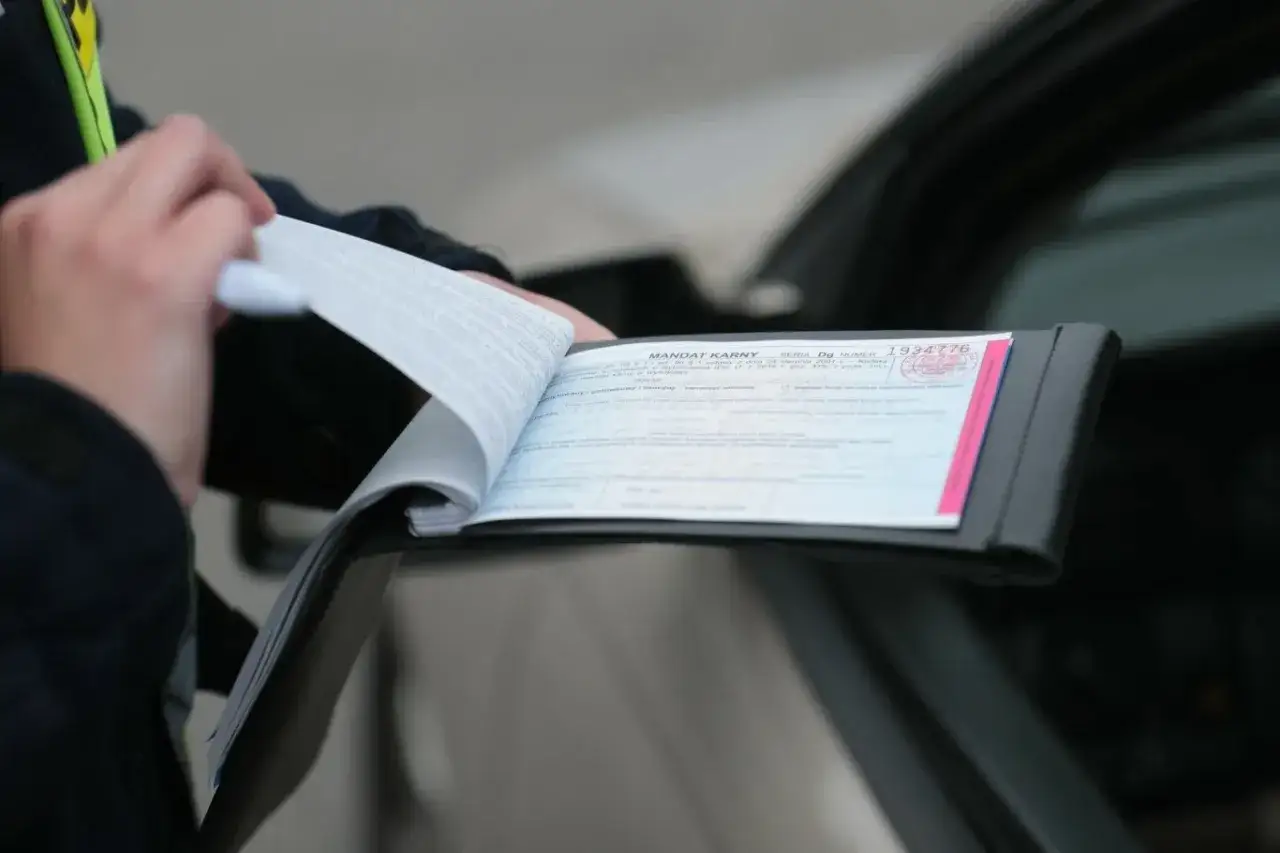 Policjant wypisuje mandat karny w notesie przy samochodzie.