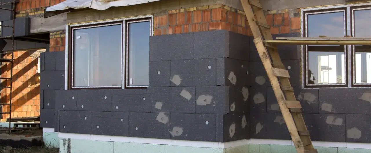 Budowa domu w toku, trwa ocieplanie ścian zewnętrznych styropianem. Widać okna i fragment rusztowania.