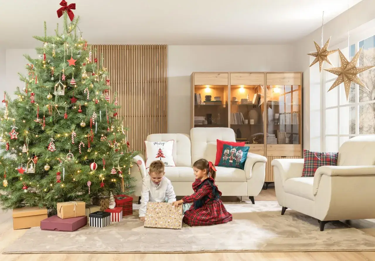 Dzieci rozpakowują prezenty przy choince na czerwono. W tle kanapa i witryna.