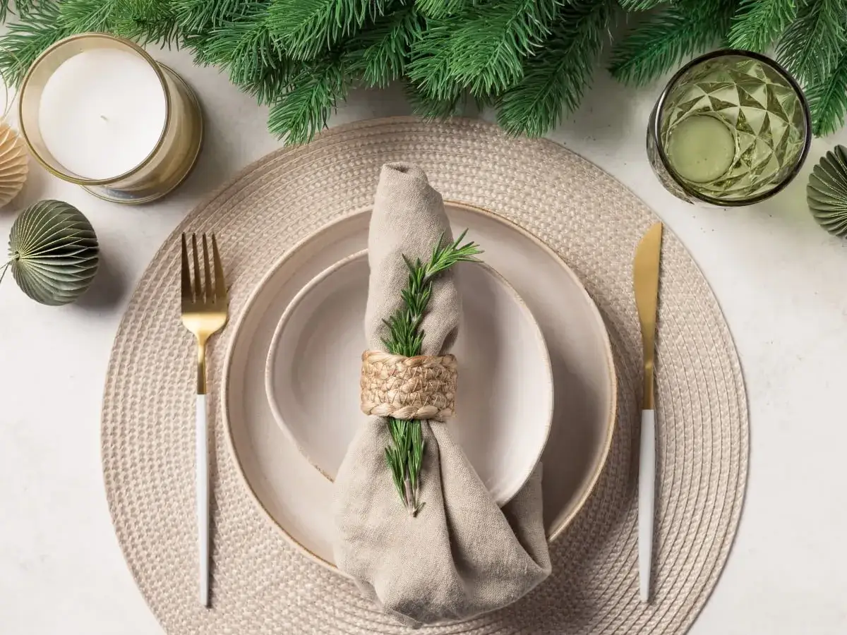 Stół nakryty do kolacji z serwetką ozdobioną rozmarynem, złotymi sztućcami i świecą.