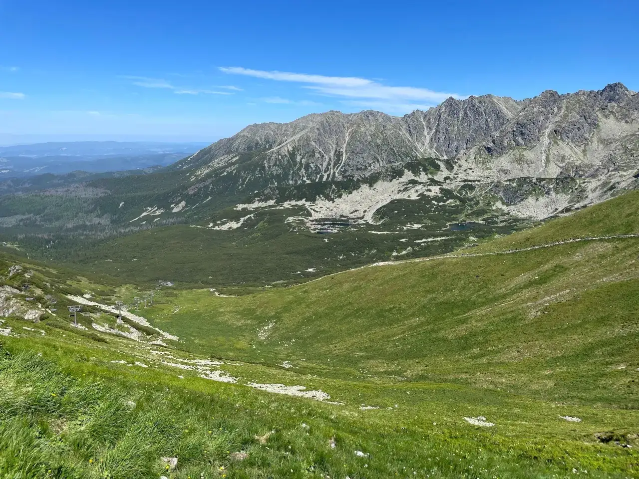 Widok na Tatry z zielonymi zboczami i skalistymi szczytami. W oddali widać budynki. Zastanawiasz się, ile km jest na Kasprowy Wierch?