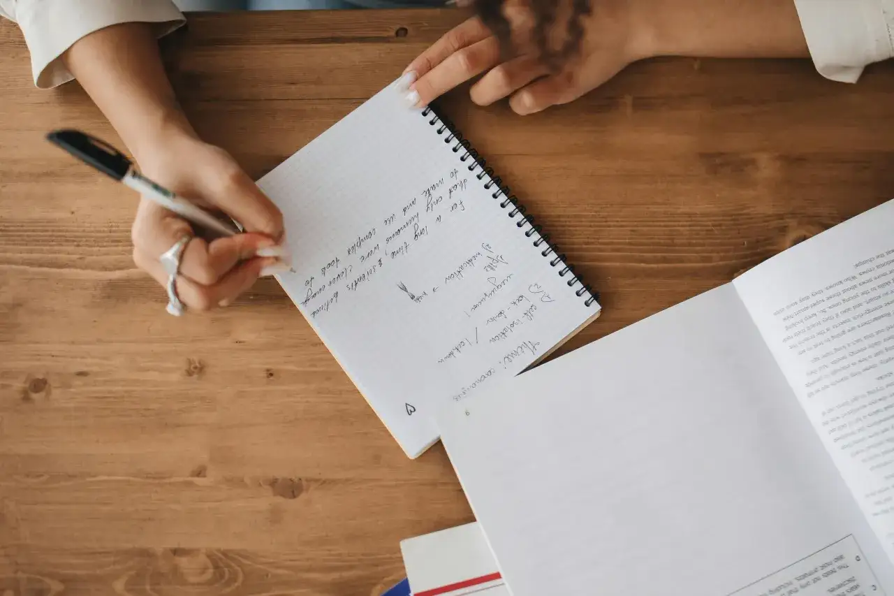 A person's hands are writing notes in a notebook. The notes mention "Theme: coronavirus", "self-isolation", and "lockdown".