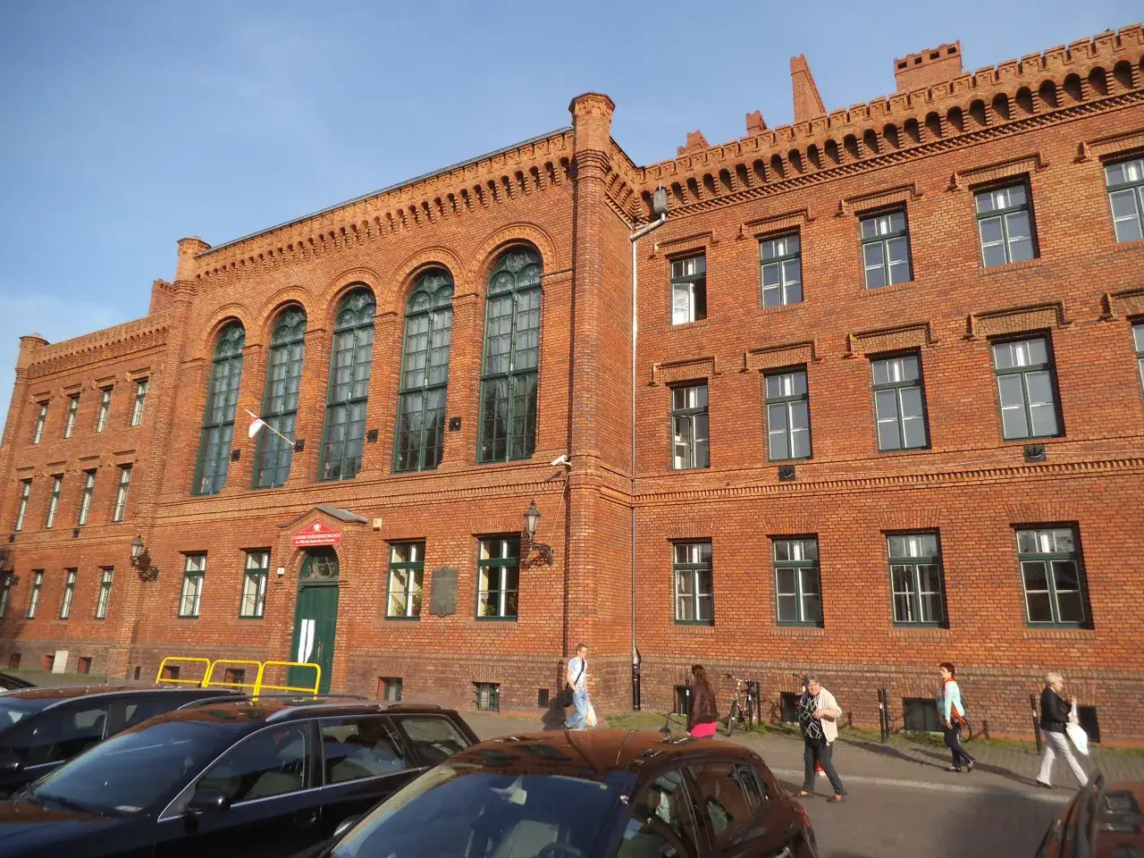 Ceglana kamienica z zielonymi oknami, gdzie w wieku 19 lat kończy się liceum. Ludzie idą chodnikiem.