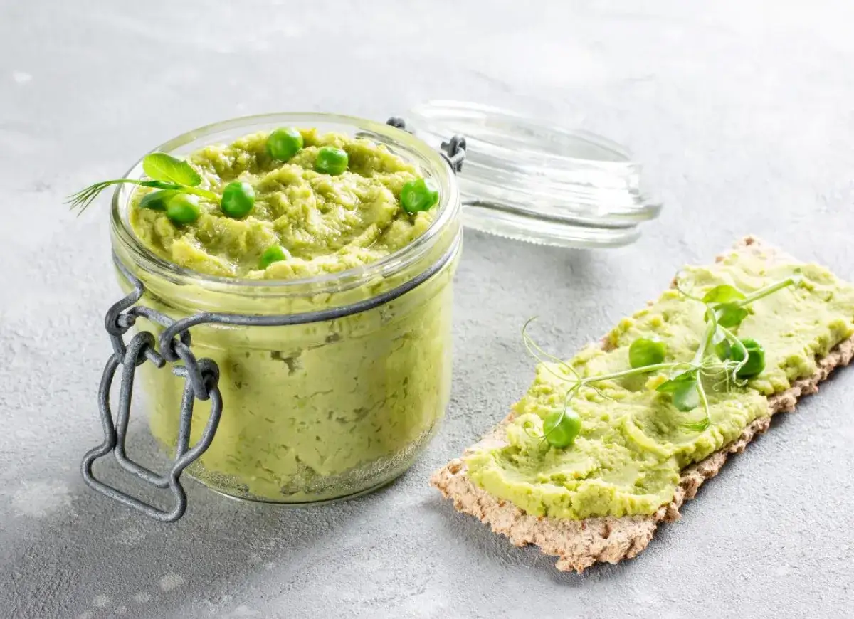 Słoik pełen pysznej, domowej pasty z zielonego groszku, obok kromka chrupkiego pieczywa z tą samą pastą i kiełkami.