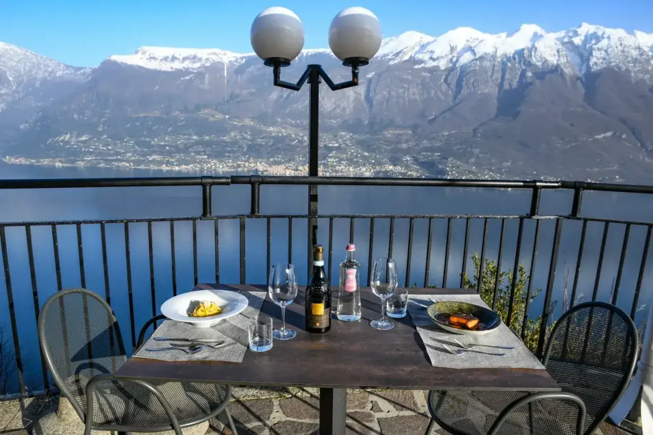 Stół z winem i jedzeniem na tarasie z widokiem na jezioro i ośnieżone góry. Idealne miejsce na romantyczną kolację z widokiem, prawdziwa "terrazza del brivido".