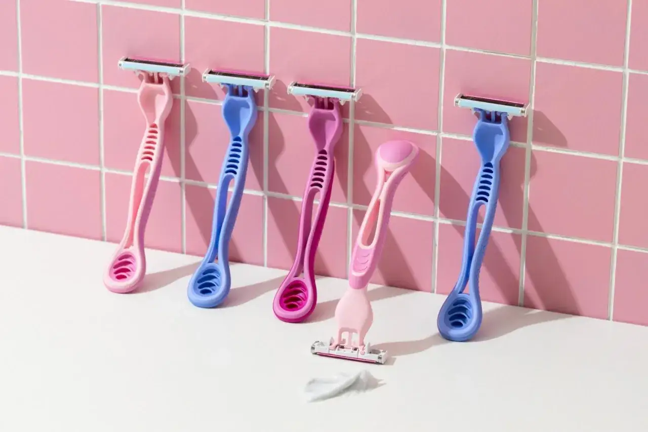 Pięć kolorowych maszynek do golenia (różowe i niebieskie) na białej powierzchni przed różową, kafelkową ścianą.