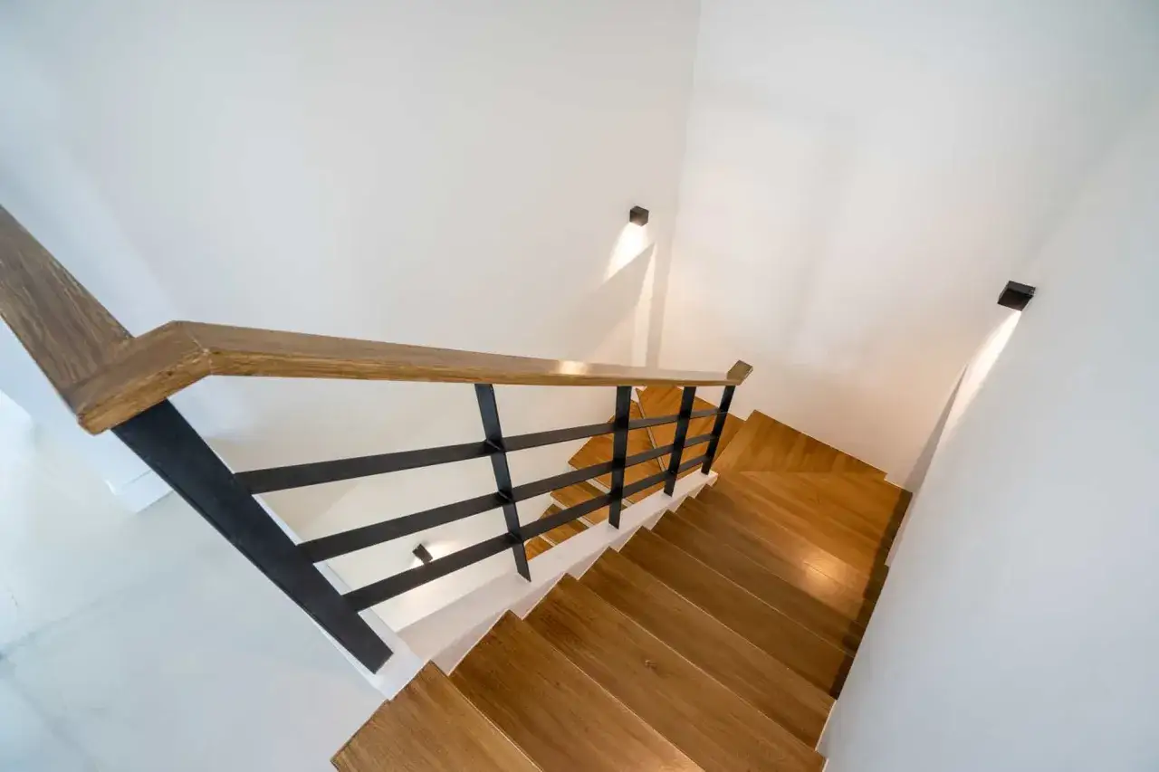 Nowoczesne schody z drewnianymi stopniami i czarną balustradą. Jak oświetlić schody w domu? Dyskretne kinkiety na ścianie tworzą nastrojowy blask.