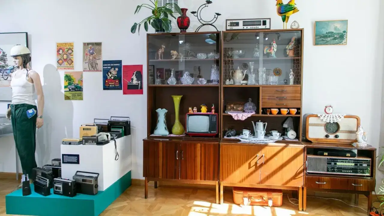 Stylizacja PRL: manekin w białej koszulce i zielonych spodniach, meblościanka z bibelotami, radia i telewizor.