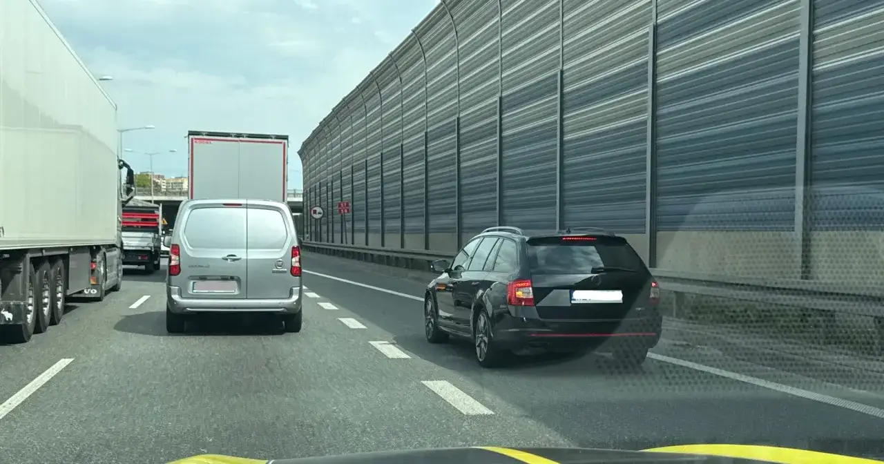 Czarny kombi jedzie pasem awaryjnym, ryzykując mandat. W tle inne pojazdy i ekrany akustyczne.
