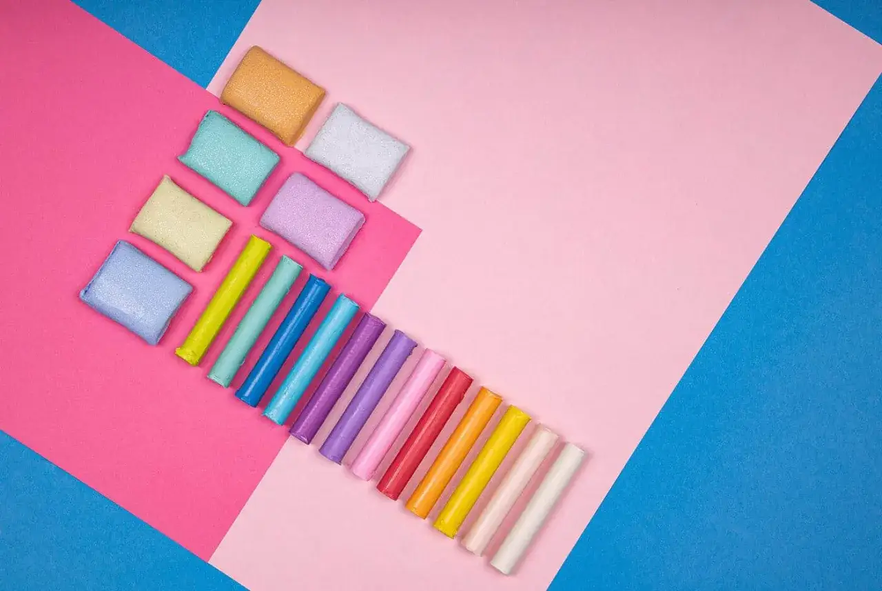 Kolorowe wałeczki i kostki modeliny, które twardnieją po wyschnięciu, ułożone na tle różowych i niebieskich kartek.