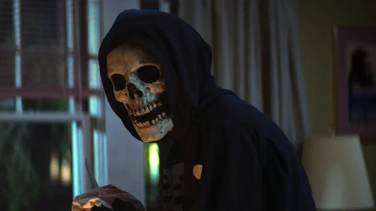 Eine Person mit Totenkopfmaske und Kapuze hält blutige Gegenstände.