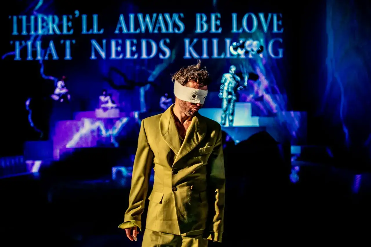 Mężczyzna w oliwkowej marynarce z zawiązanymi oczami na scenie. W tle napisy "THERE'LL ALWAYS BE LOVE THAT NEEDS KILLING". Spektakl w Warszawie.