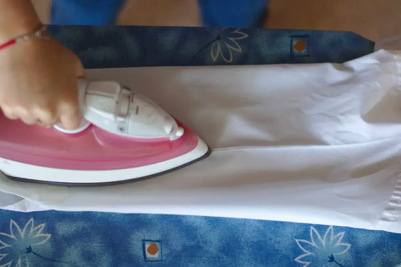 Dłoń prasuje biały rękaw koszuli różowym żelazkiem. Widać fragment deski do prasowania z niebieskim wzorem.