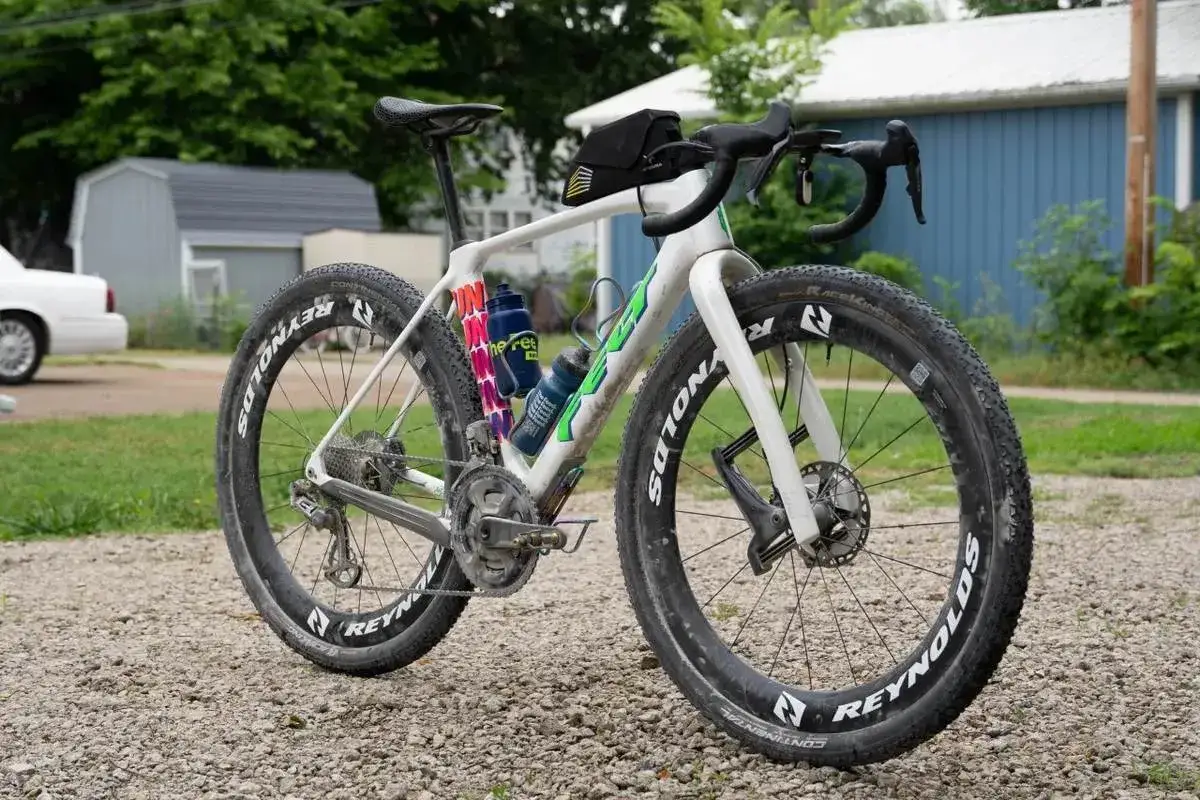 Biały rower gravelowy z szerokimi oponami i sakiewką na kierownicy, gotowy na przygodę. Czy to gravel czy szosa, zależy od terenu!