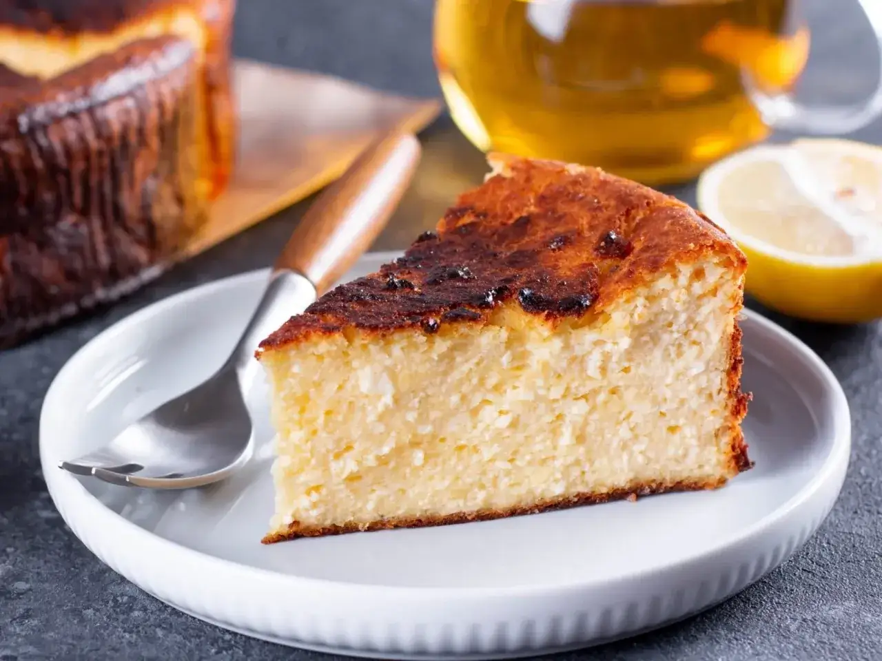 Pyszny sernik, który nie opada na herbatnikach, z lekko przypieczoną skórką, podany na talerzyku z widelcem. Obok plaster cytryny i filiżanka herbaty.