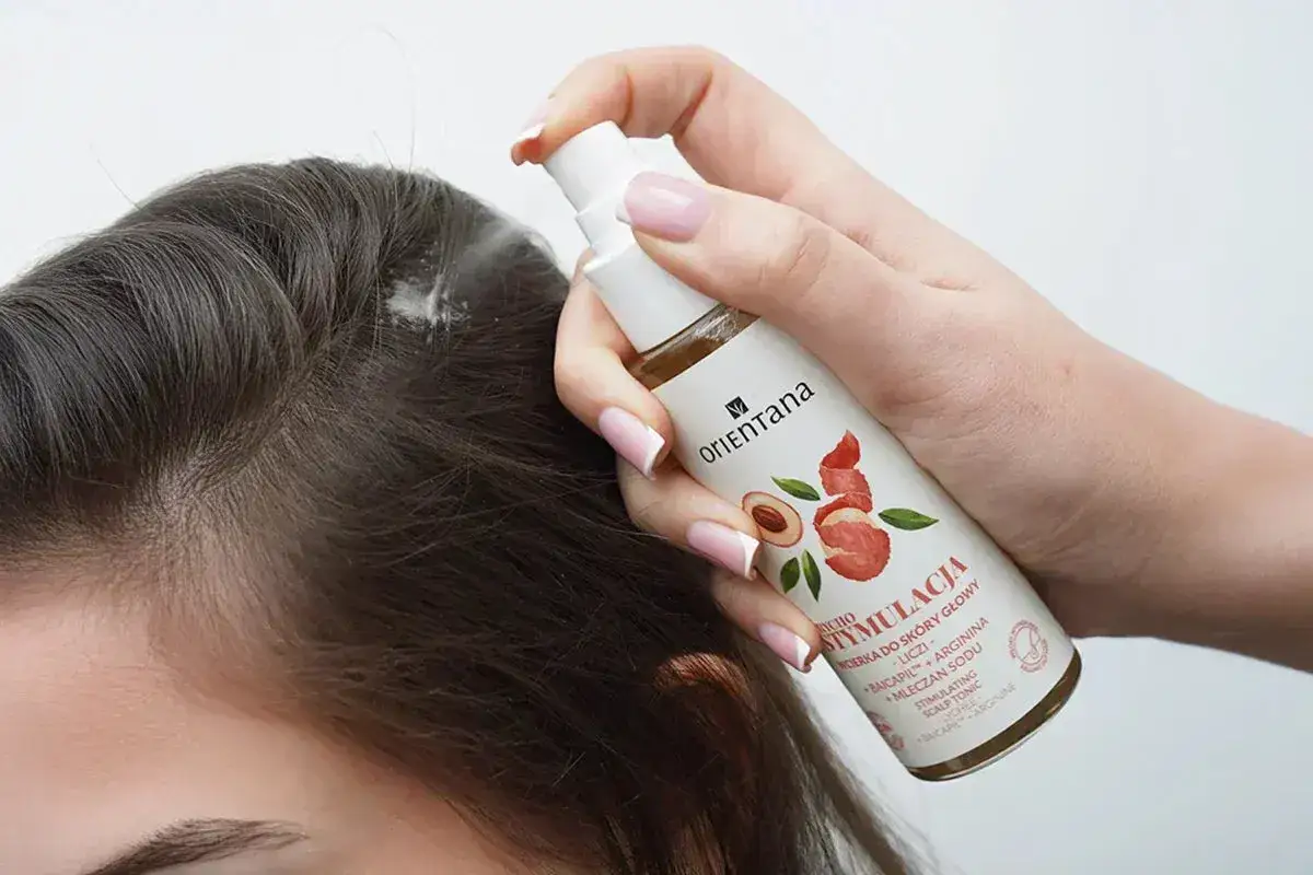 Dłoń aplikuje wcierkę do skóry głowy z butelki z atomizerem.