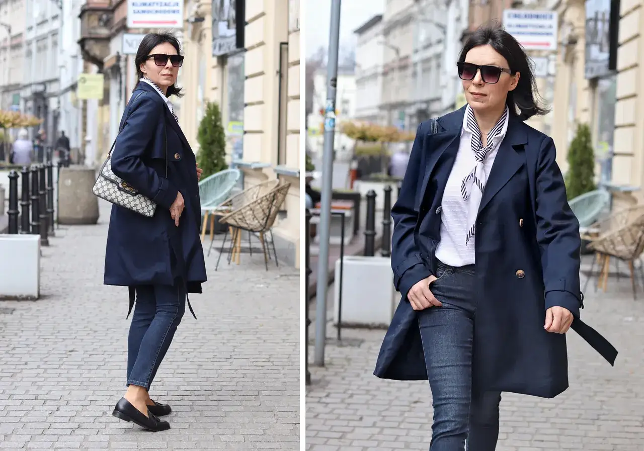 Elegancka kobieta w granatowym trenczu damskim, białej bluzce w paski i jeansach spaceruje po brukowanej ulicy.