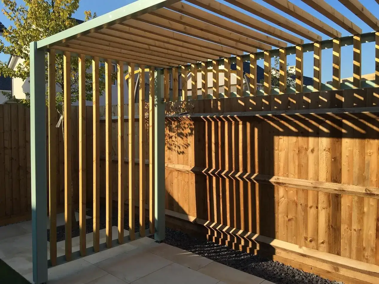 Nowa pergola z drewnianymi listwami i metalową konstrukcją, z cieniami na drewnianym płocie.