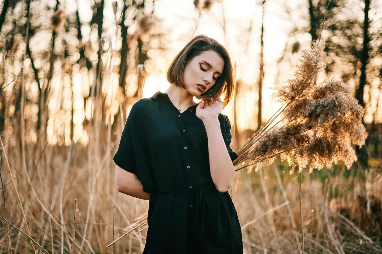 Młoda kobieta w czarnej sukience pozuje na tle zachodzącego słońca i trzcin. Idealna modelka do nauki kurs fotografii portretowej.
