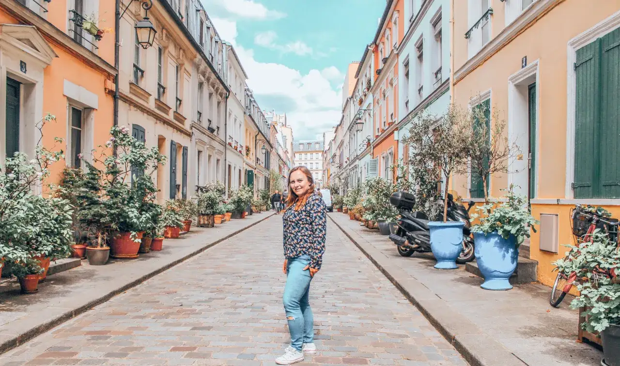 Urocza ulica w 16. dzielnicy Paryża, z kolorowymi kamienicami, mnóstwem zieleni w donicach i uśmiechniętą kobietą.