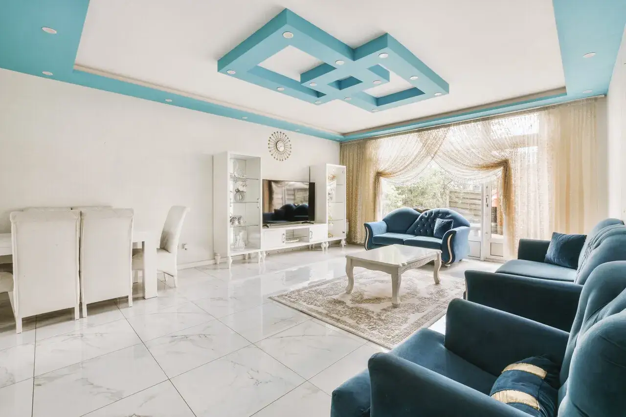 Nowoczesny salon z błękitnym, geometrycznym, dekoracyjnym malowidłem na suficie. Wygodne, granatowe sofy i białe meble tworzą elegancką przestrzeń.