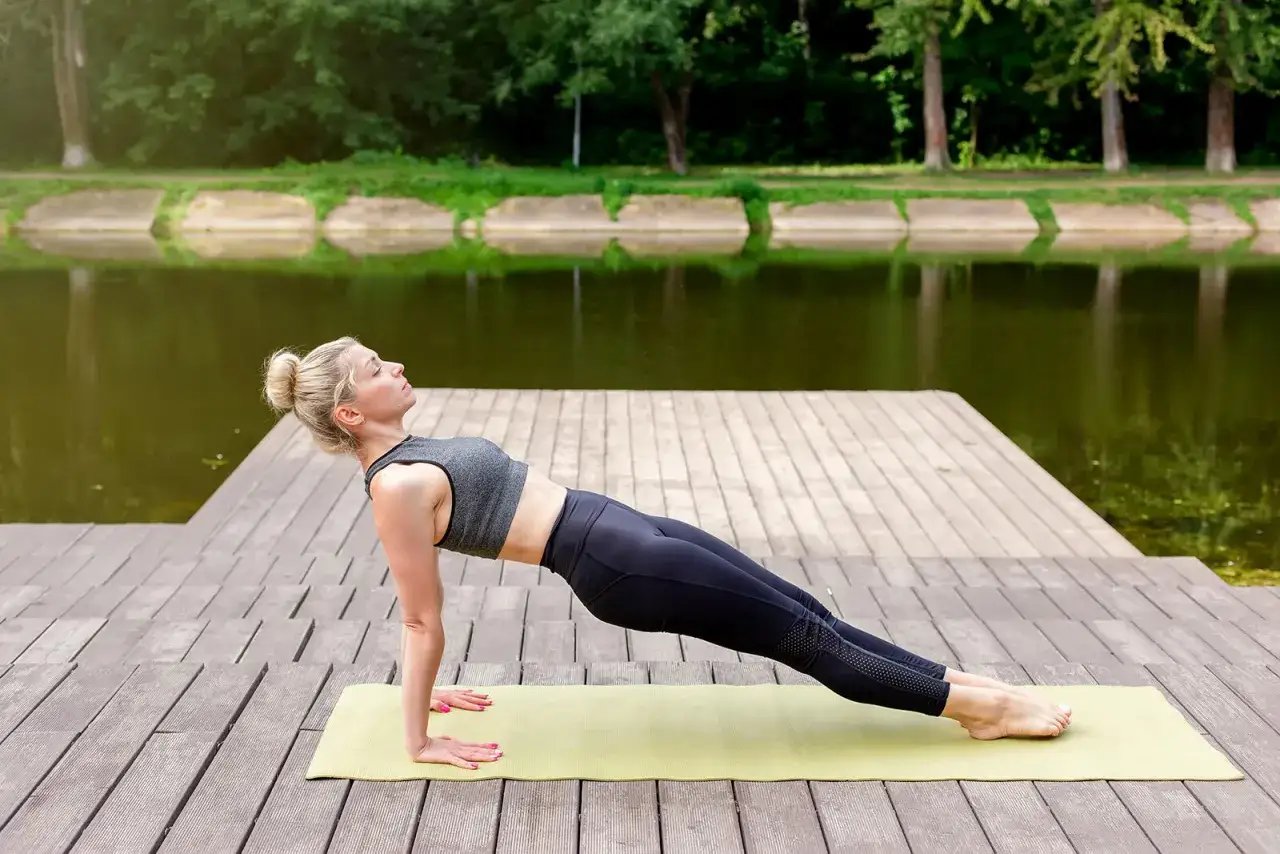 Kobieta wykonuje plank tyłem na macie nad jeziorem, wzmacniając mięśnie.