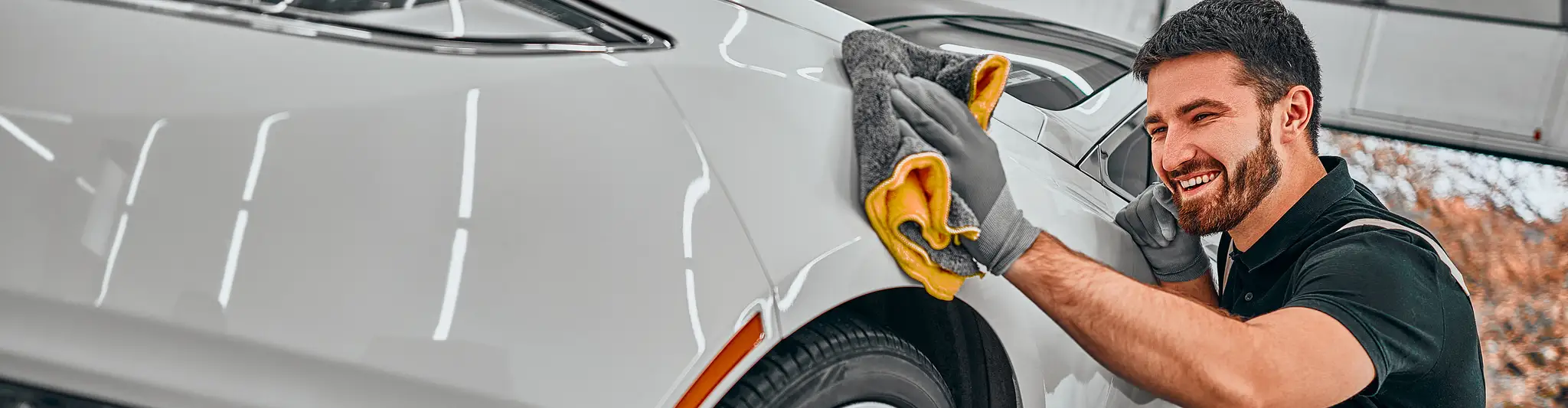 Uśmiechnięty mężczyzna w rękawiczkach poleruje białe auto szarą mikrofibrą z żółtym obszyciem.