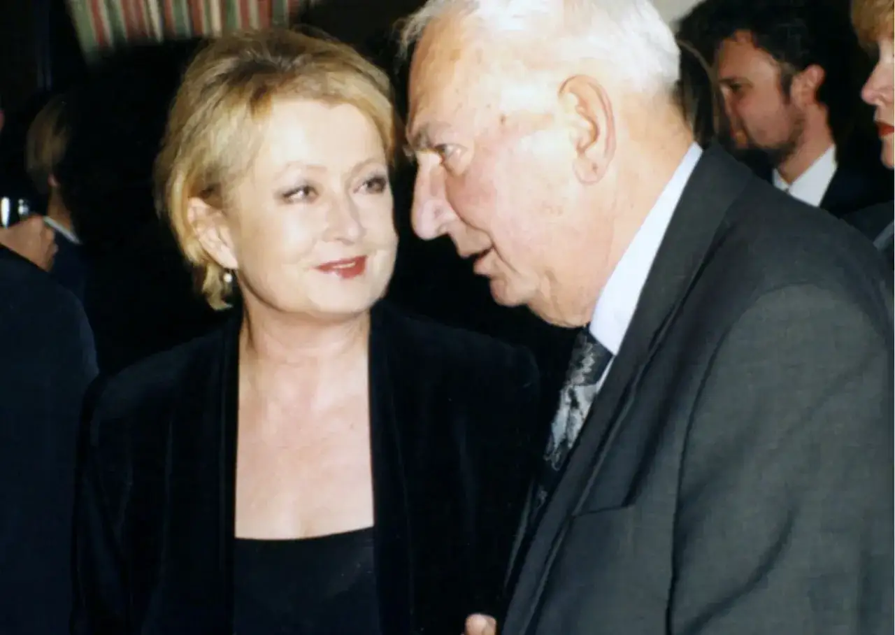 Ewa Holoubek w czarnej sukni i marynarce rozmawia z mężczyzną w garniturze.