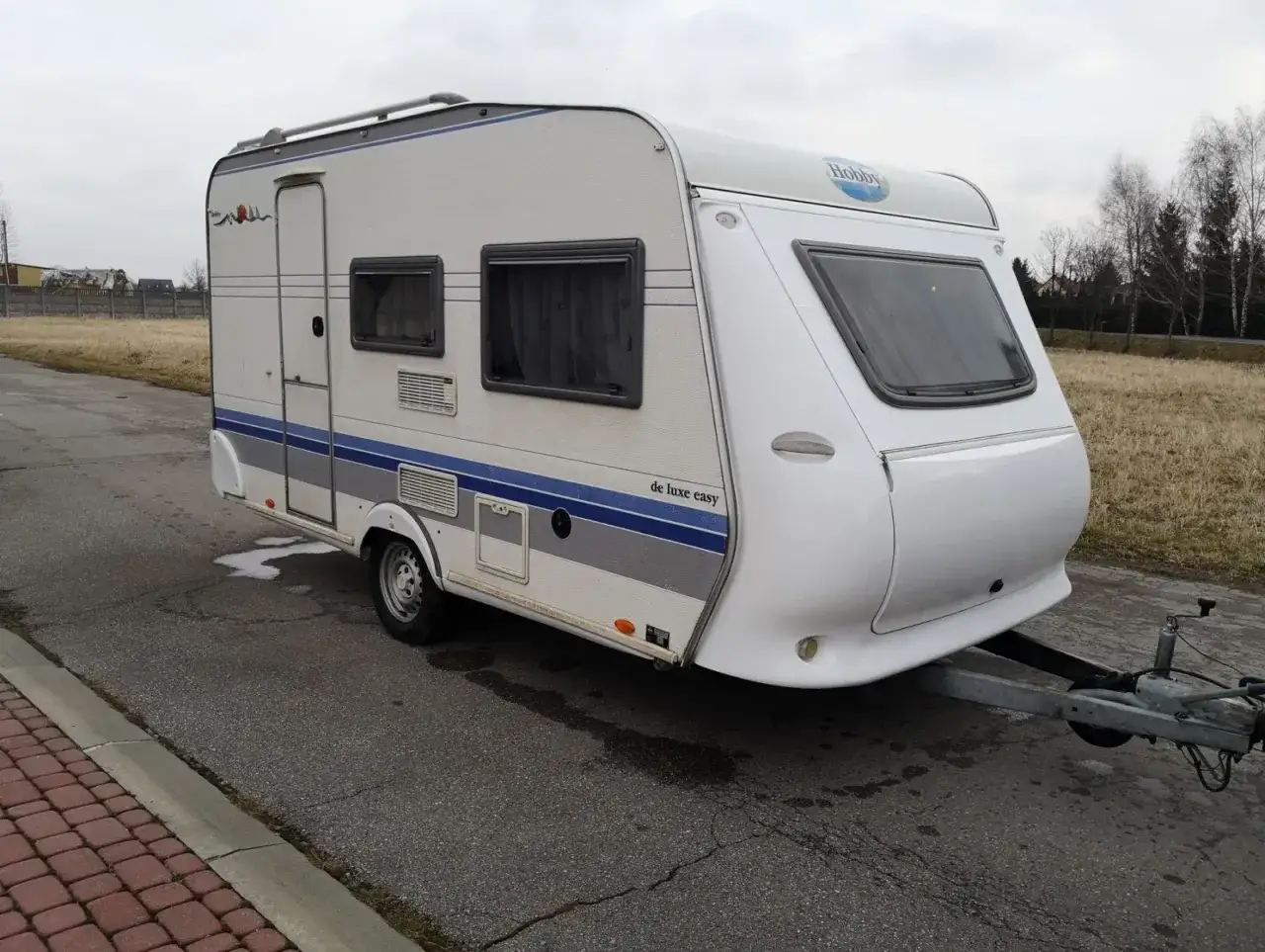 Biała przyczepa kempingowa Hobby 400 de luxe easy z niebieskimi pasami, gotowa na wakacyjne podróże.