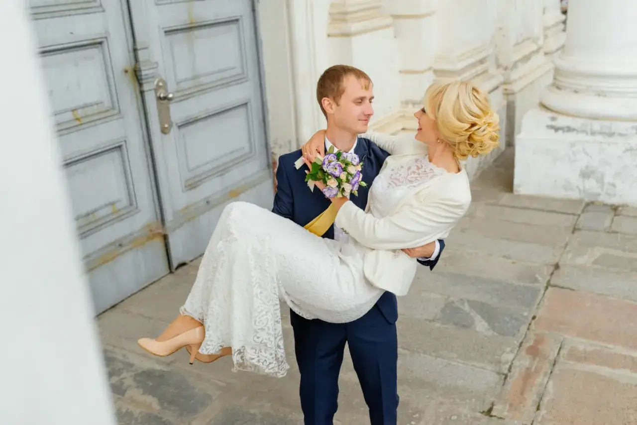 Pan młody niesie pannę młodą na rękach. Zastanawiasz się nad skutkami spóźnienia się z rezerwacją fotografa? Dlaczego 18 miesięcy to minimum?