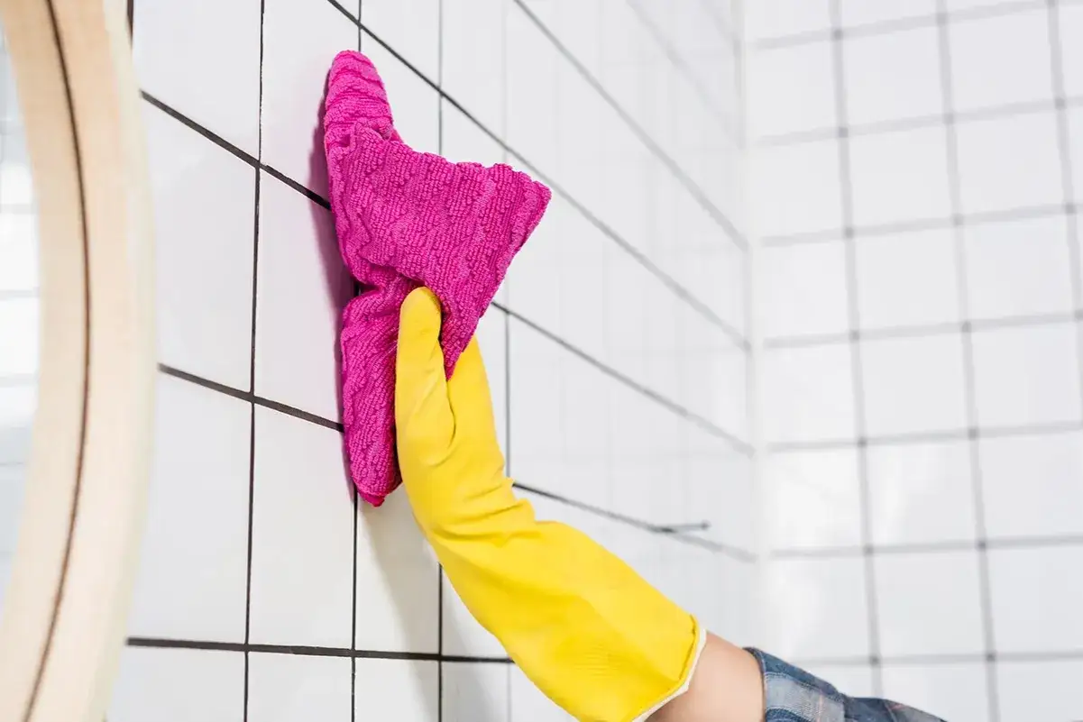 Dłoń w żółtej rękawiczce czyści różową ściereczką białe płytki.