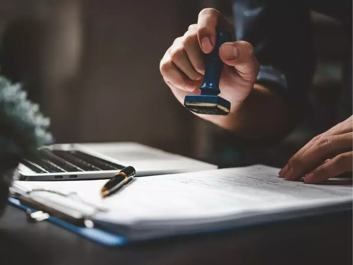 Dłoń z pieczęcią nad dokumentem, symbolizującym notarialne poświadczenie dziedziczenia. Obok laptop i długopis.