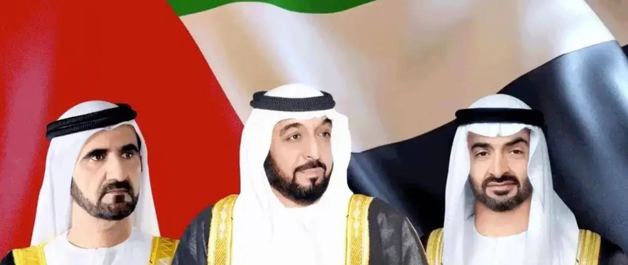 Trzech arabskich przywódców w tradycyjnych strojach, na tle flagi Zjednoczonych Emiratów Arabskich. Jeden z nich, być może najbogatszy szejk na świecie, patrzy wprost.