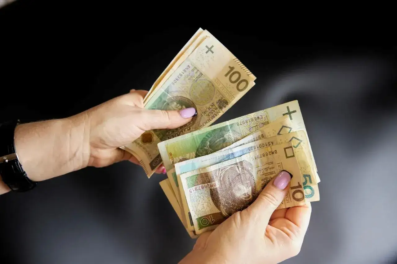Dwie dłonie liczą polskie banknoty. Jedna trzyma 100 zł, druga 10 i 50 zł. Może to symbolizować odszkodowania wojenne dla spadkobierców.