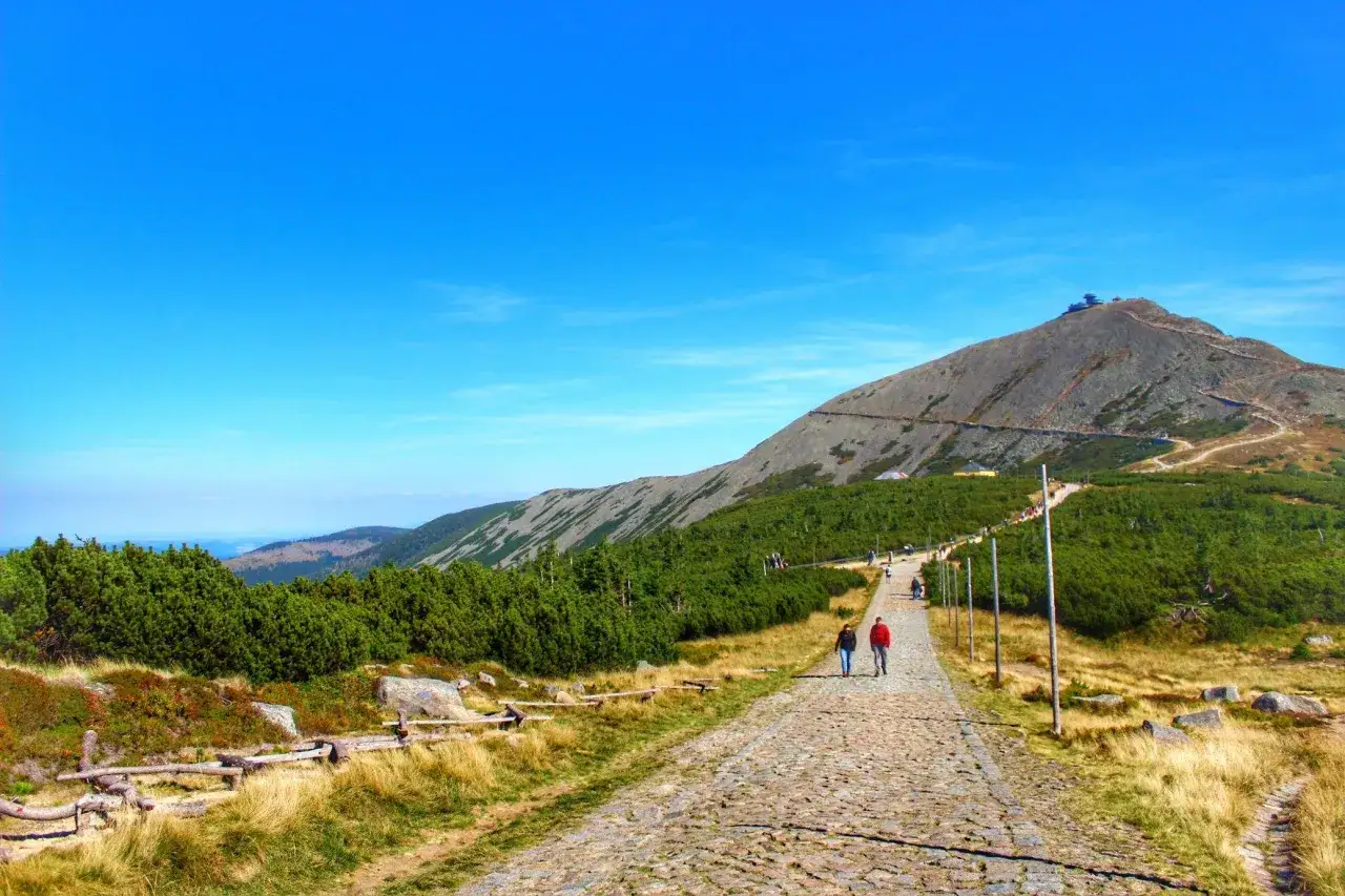 Kamienista ścieżka prowadzi na szczyt Śnieżki. Turyści idą w kierunku górskiej budowli.