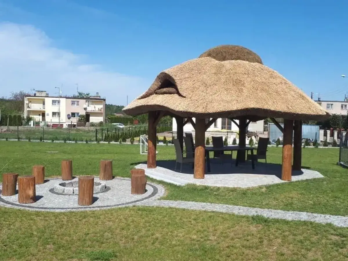 Altana z bali ze słomianym dachem, stołem i krzesłami w środku, otoczona zielenią i kamiennym kręgiem z pni.