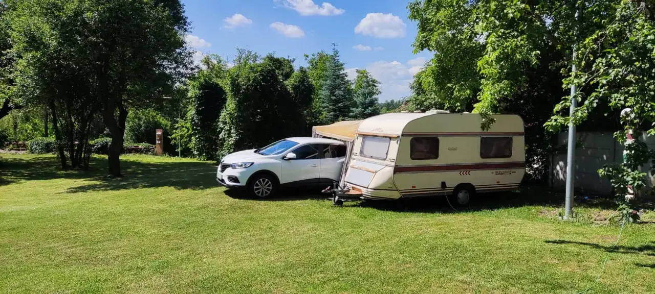 Biały samochód i przyczepa kempingowa na zielonej trawie, otoczone drzewami. Idealne miejsce na **camping smok**.