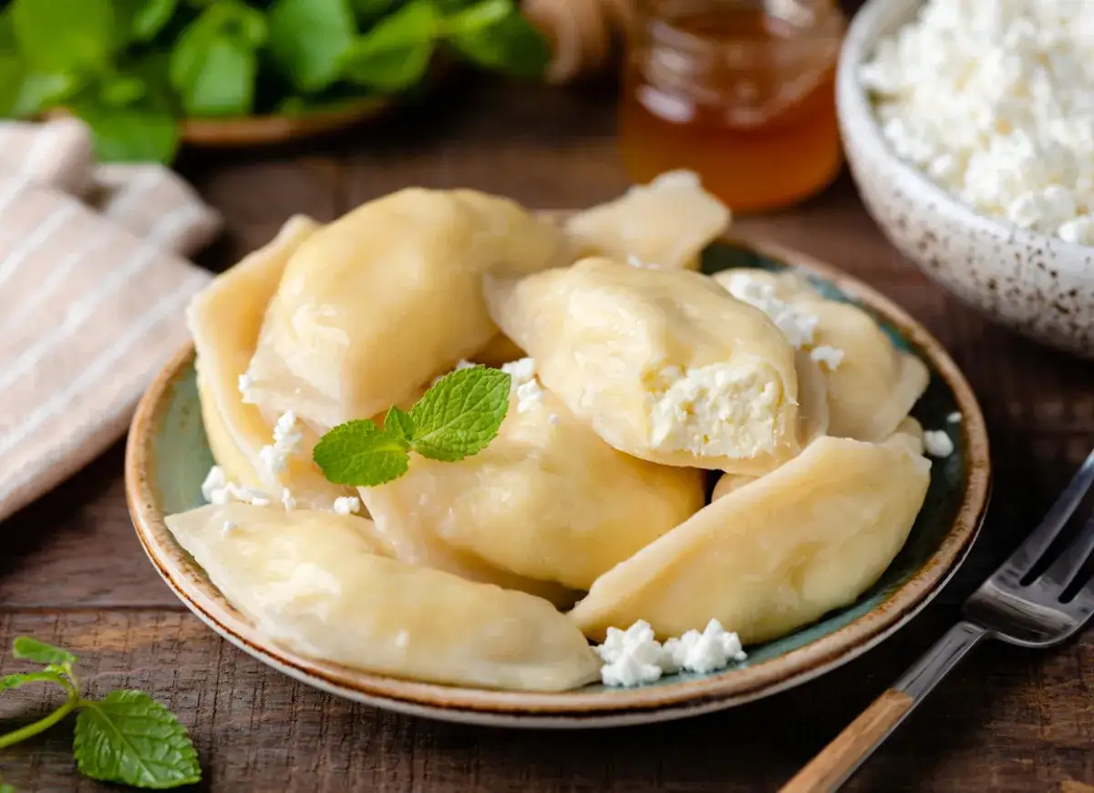 Pyszne pierogi z serem, udekorowane miętą i posypane białym serem. Obok słoiczek miodu i miseczka z serem.