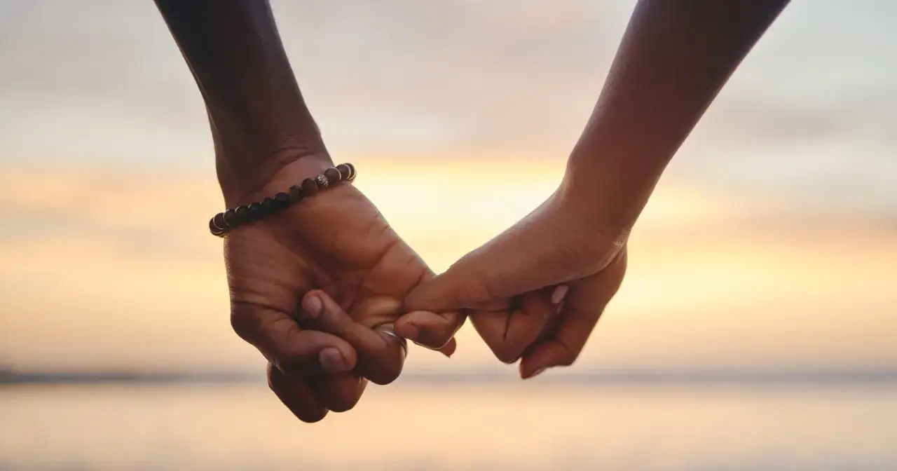 Dłonie splecione na tle zachodzącego słońca, budzące tęsknotę za pierwszą miłością.