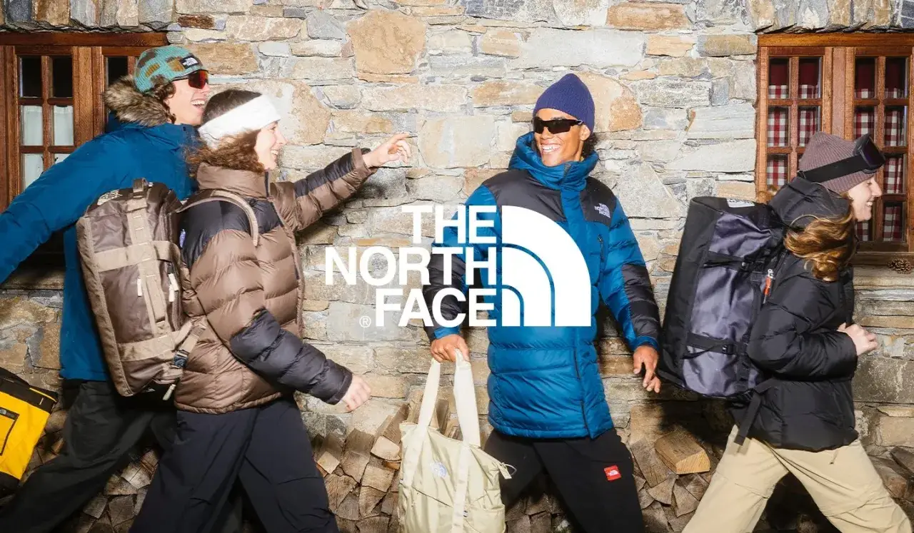 Cztery osoby w kurtkach The North Face, z plecakami i torbami, na tle kamiennej ściany. Jak rozpoznać podróbkę? Sprawdź detale!