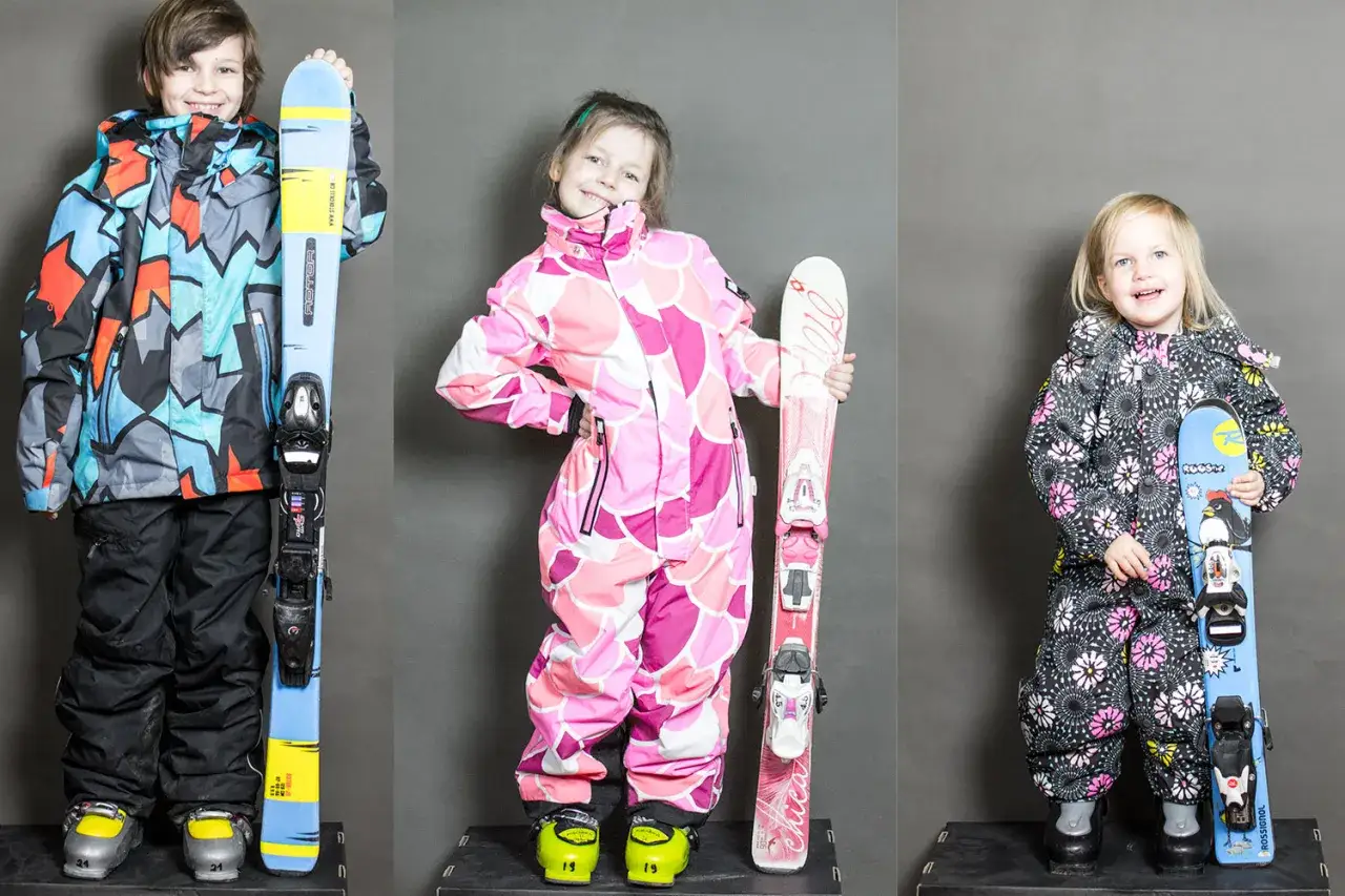 Trójka dzieci gotowa na stok. Chłopiec w kolorowej kurtce i dziewczynki w różowych i czarnych kombinezonach, każde z nartami. Idealne narty dla 3 latka i starszych dzieci.
