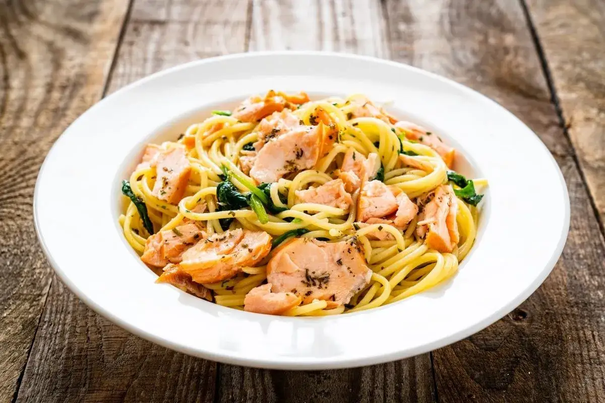 Makaron spaghetti z łososiem i szpinakiem na białym talerzu, na drewnianym tle.