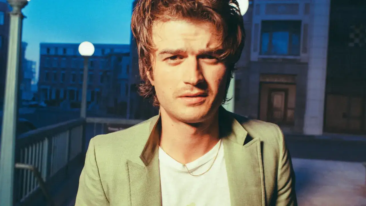 Joe Keery w jasnej marynarce, z naszyjnikiem, na tle nocnego miasta.